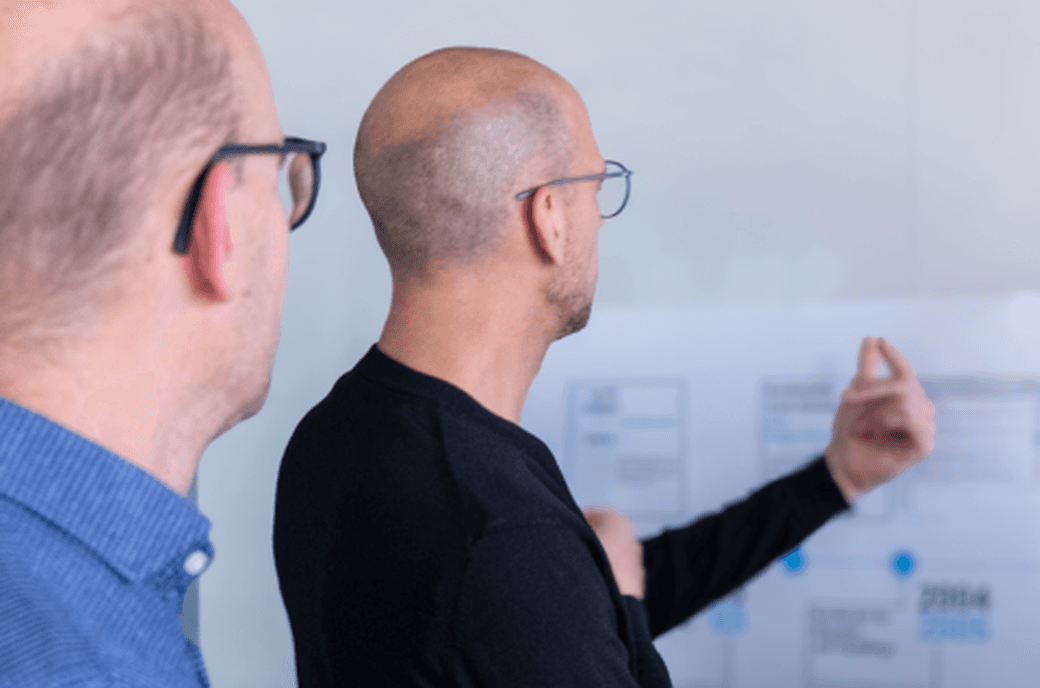 Zwei Männer stehen vor einem Whiteboard. Einer der beiden macht eine erklärende Geste